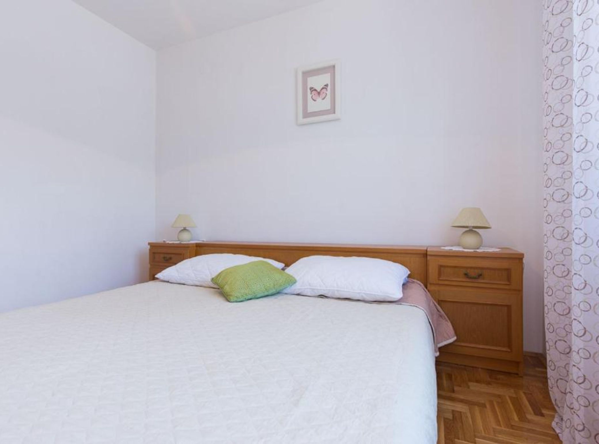 Un dormitorio con una cama, dos mesitas de noche con lámparas, un cuadro enmarcado en la pared y una cortina estampada.