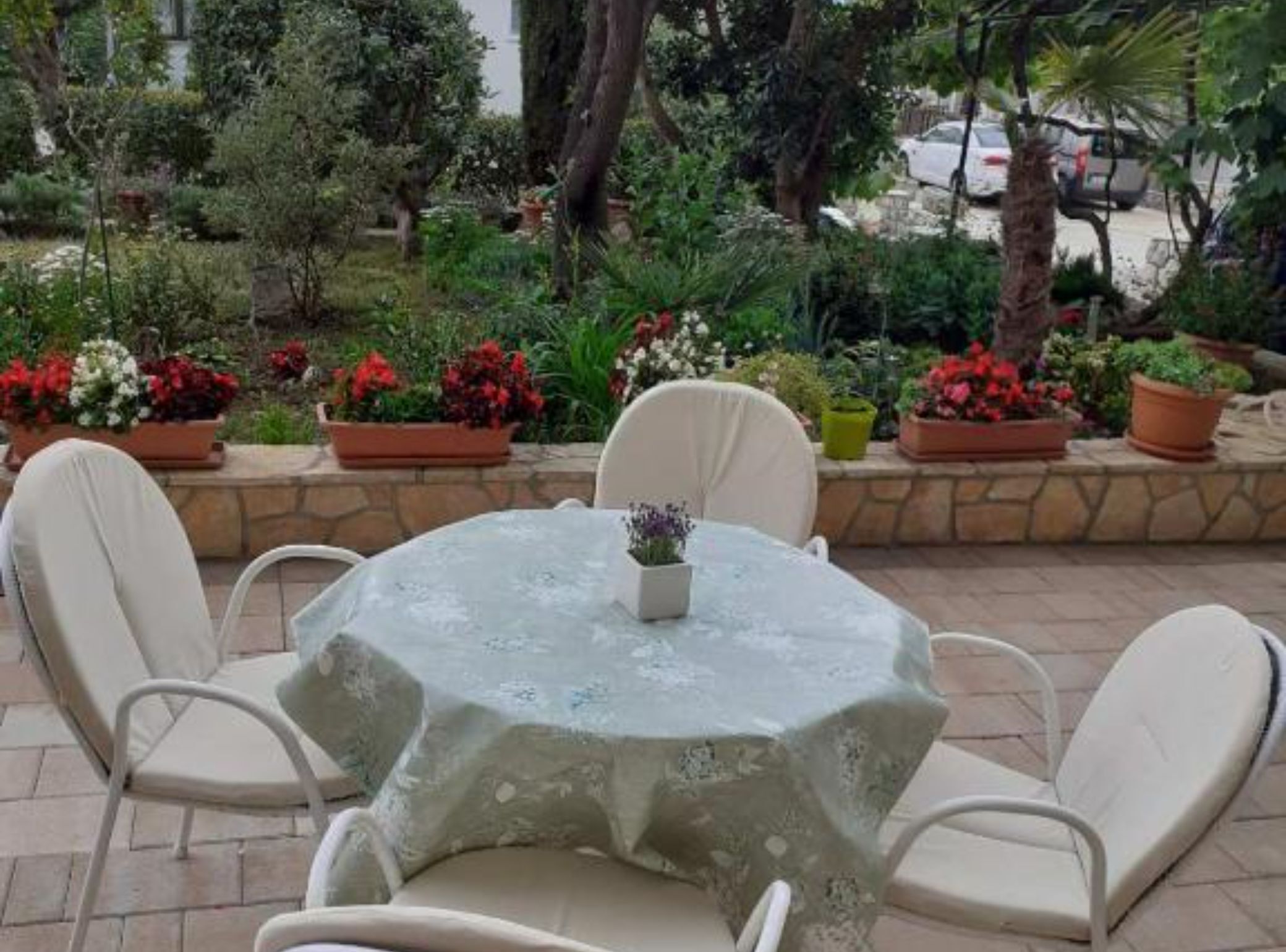 Un patio exterior con una mesa redonda y cuatro sillas, rodeado de macetas con plantas y flores.
