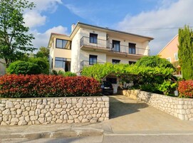 A two-story residential building with a stone wall in the foreground, a paved driveway, and a car parked in the driveway.