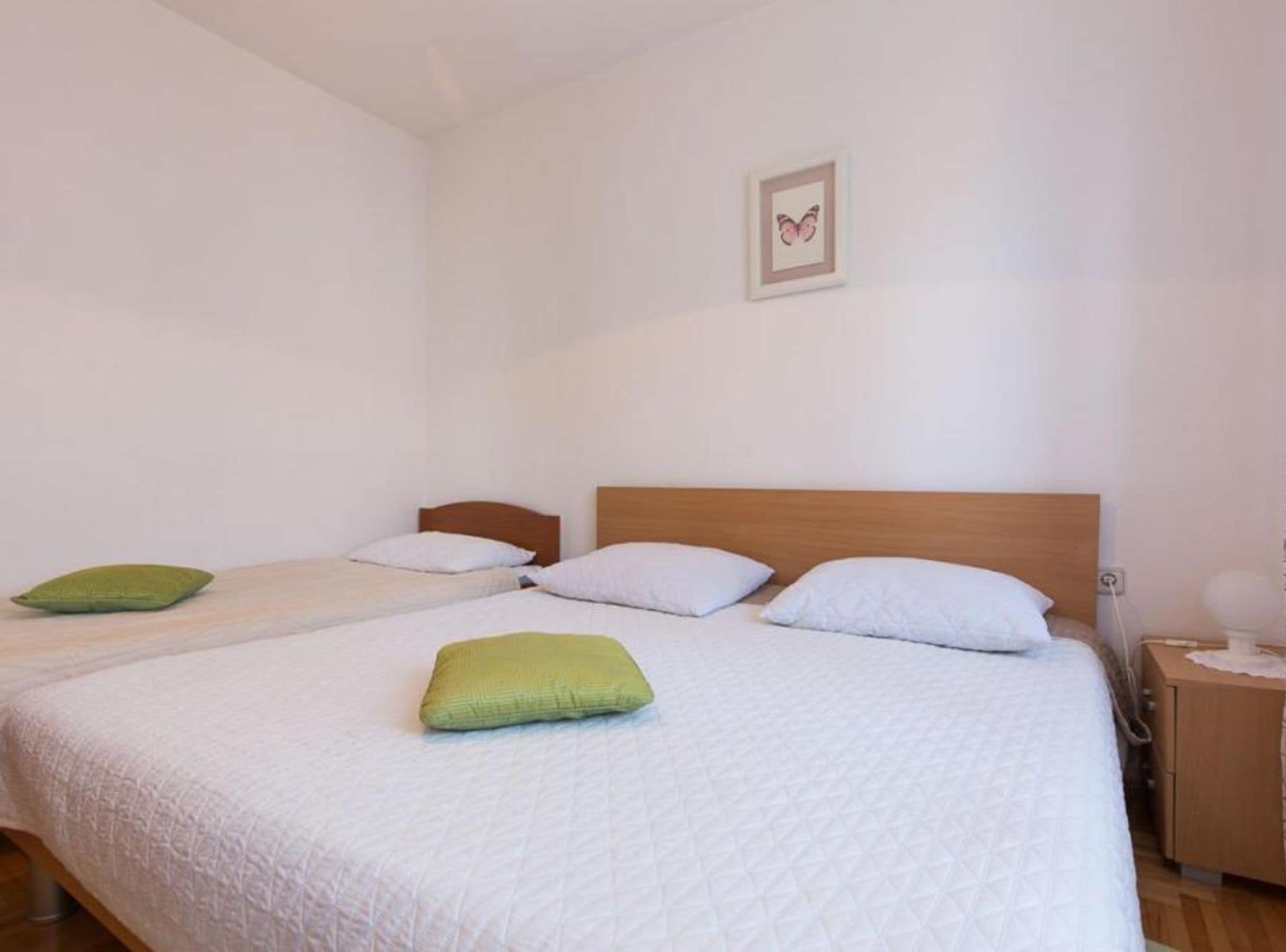 A hotel room with two single beds, each with a wooden headboard and white bedding. There is a small bedside table with a lamp on the right side.
