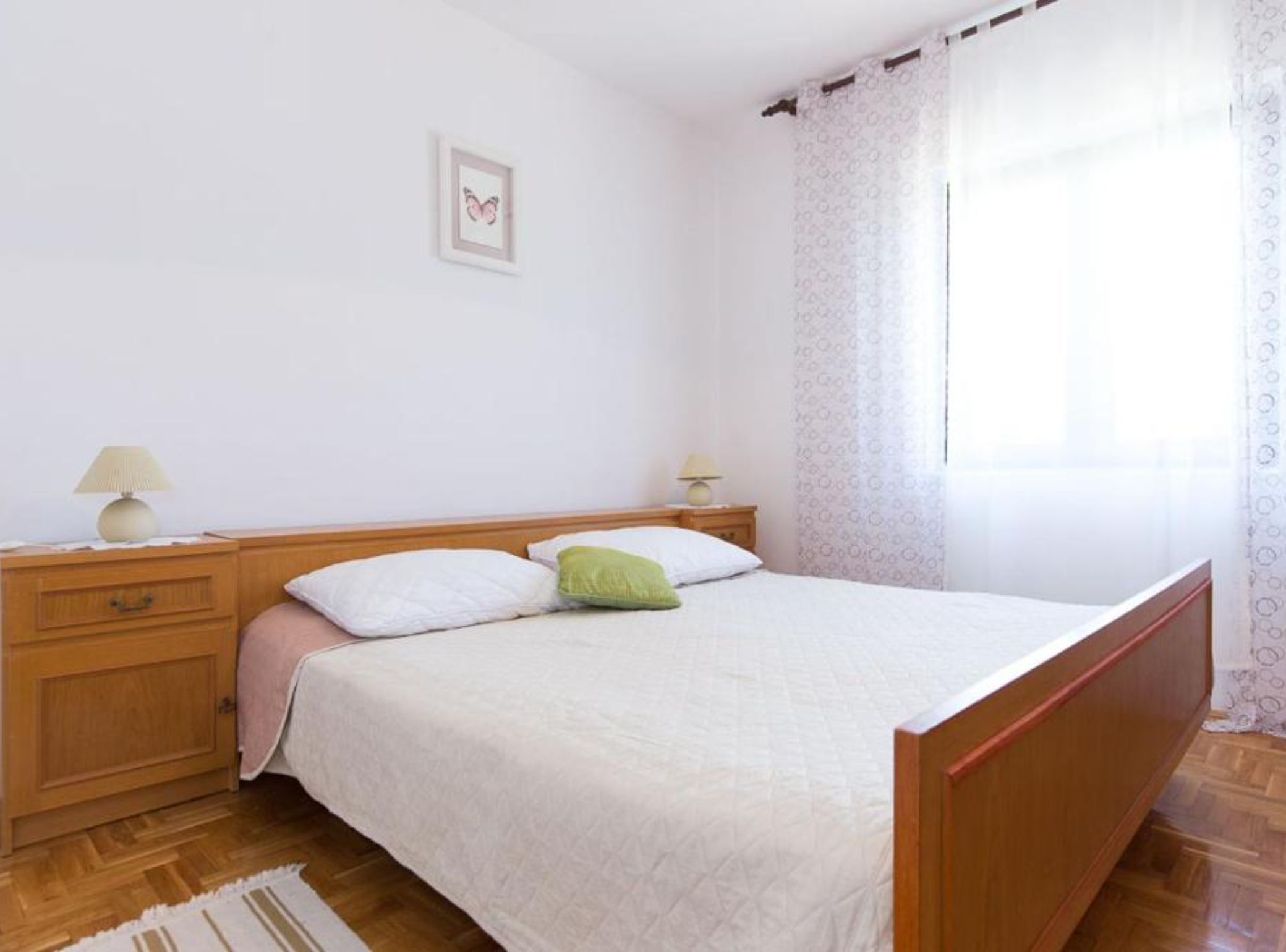 A bedroom with a bed, two bedside tables, a nightstand, a lamp, a framed picture on the wall, and a window with curtains.
