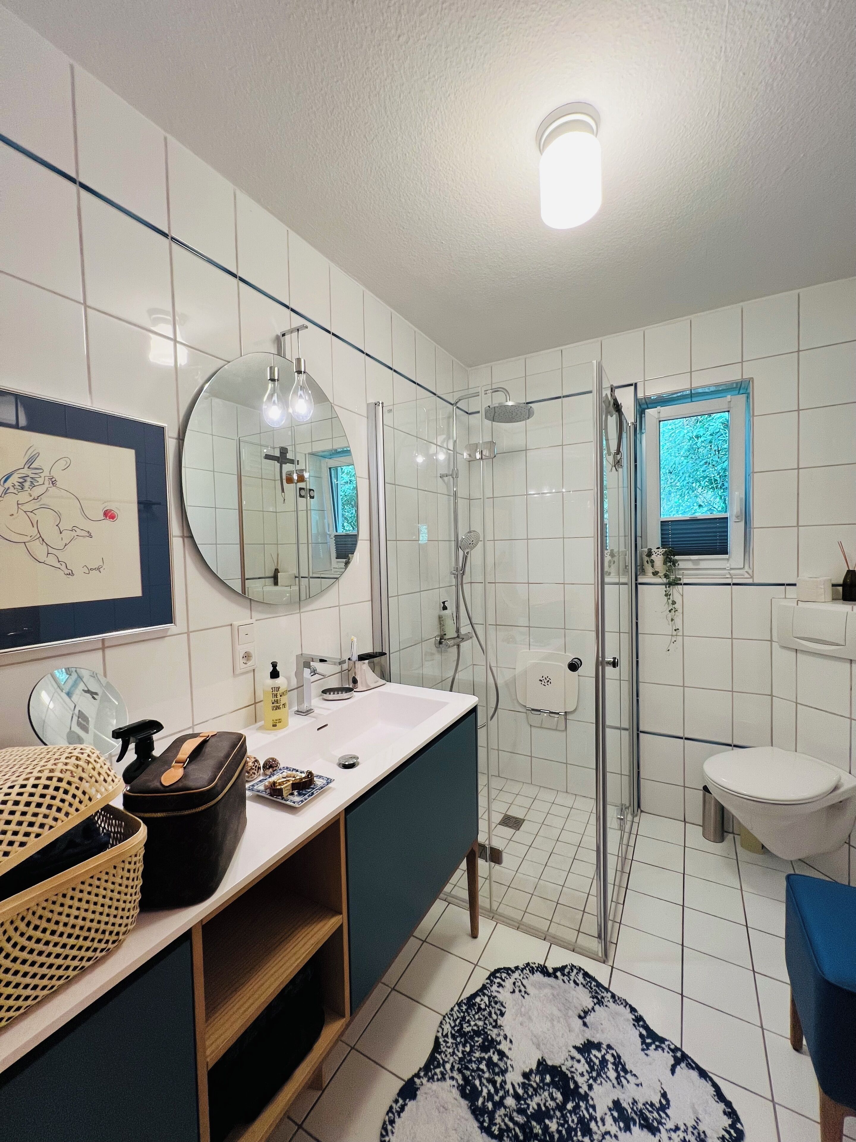 Ein modernes Badezimmer mit Dusche, Waschbecken und Toilette. Ein Spiegel, eine Leuchte und ein Fenster mit Blick auf Bäume sind vorhanden.