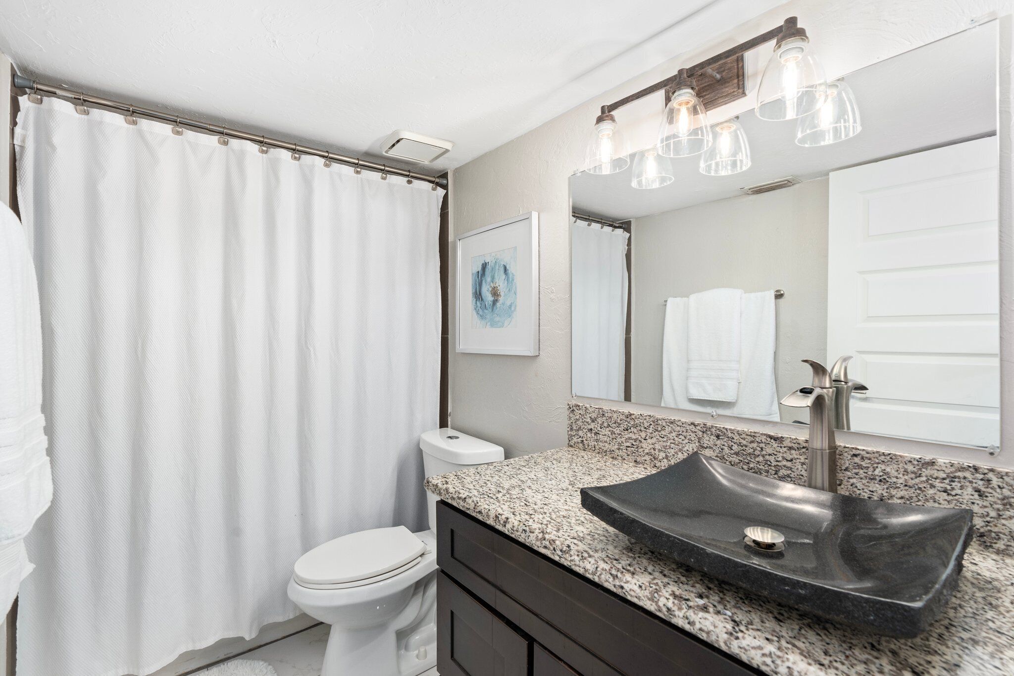 A bathroom with a white toilet, a black sink with a single basin, a shower curtain, and a mirror with a towel hanging on it.