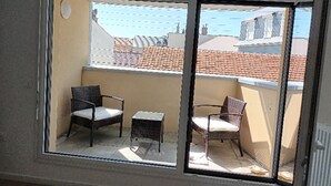 A balcony with two wicker chairs and a small table, overlooking a cityscape with red-tiled roofs.