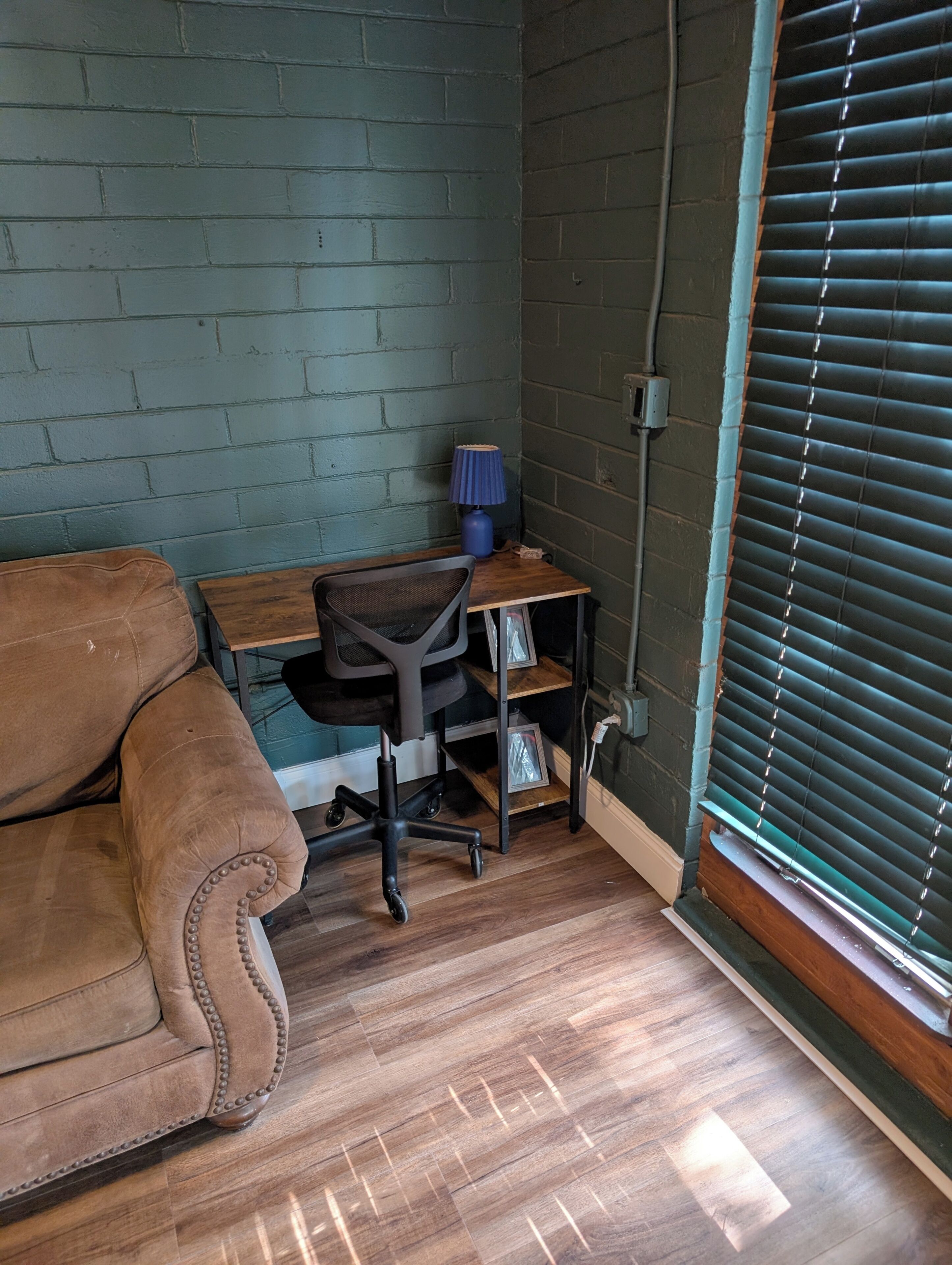 A room with a brown leather sofa, a wooden desk with a chair, a lamp, and a window with blinds.