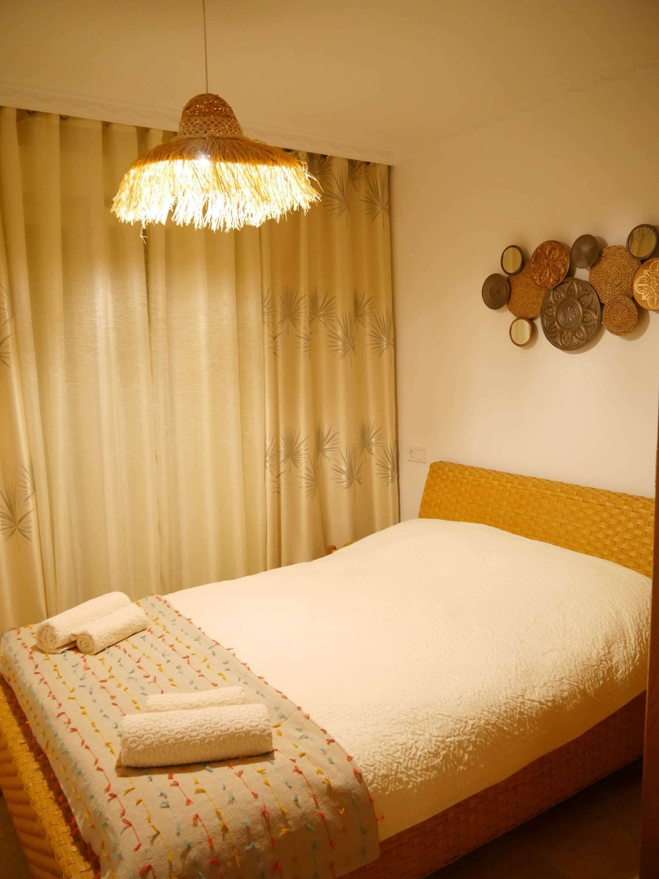 Ein ordentlich bezogenes Bett mit weißer Daunendecke und farbenfroher Decke, eine Hängelampe und eine Wand mit dekorativen Tellern.