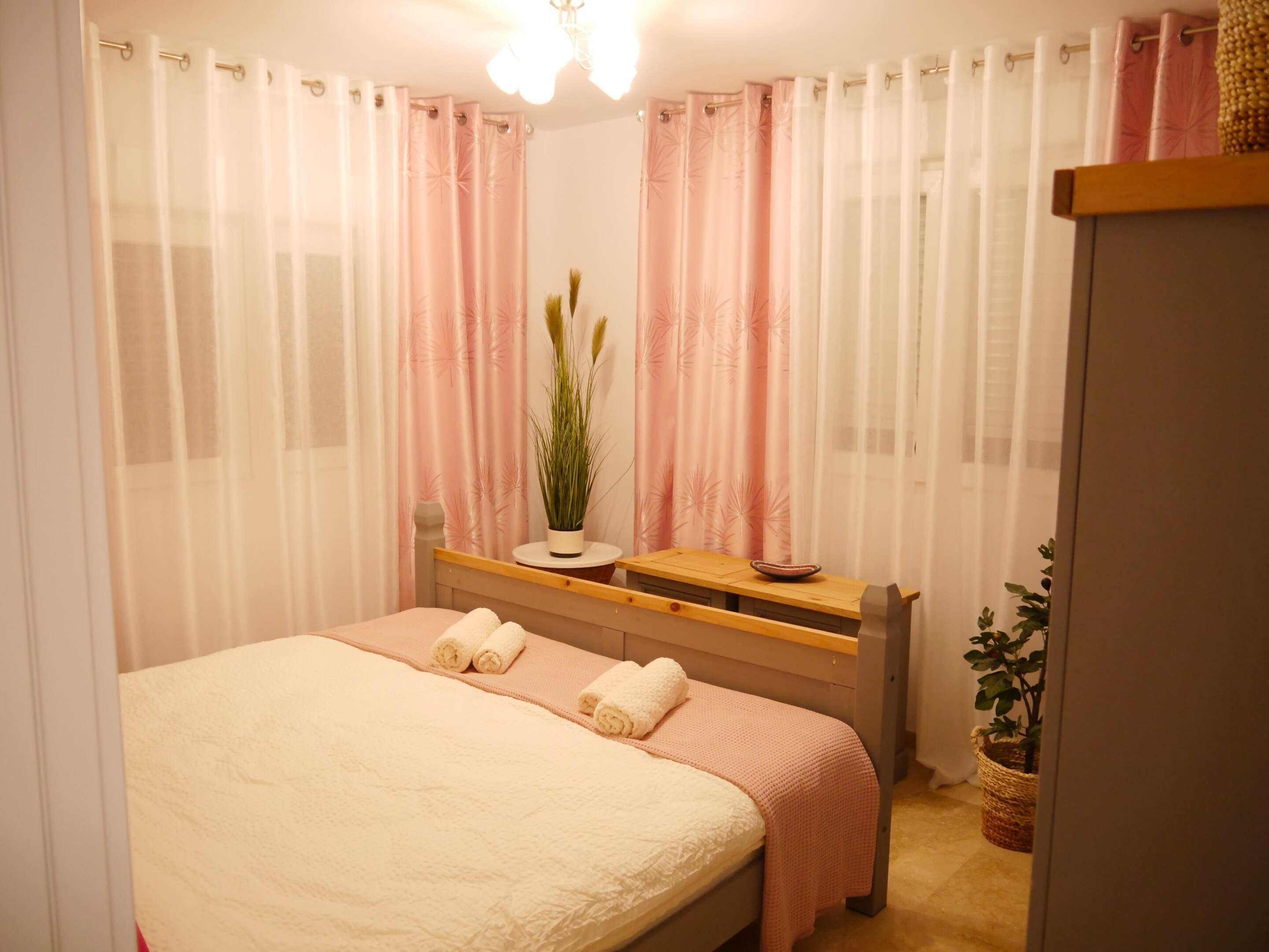 Ein Schlafzimmer mit Bett, Nachttisch, Blumentopf und Fenster mit durchsichtigen Vorhängen.