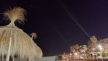 Een strand met rieten parasols, palmbomen en een verlichte boulevard.