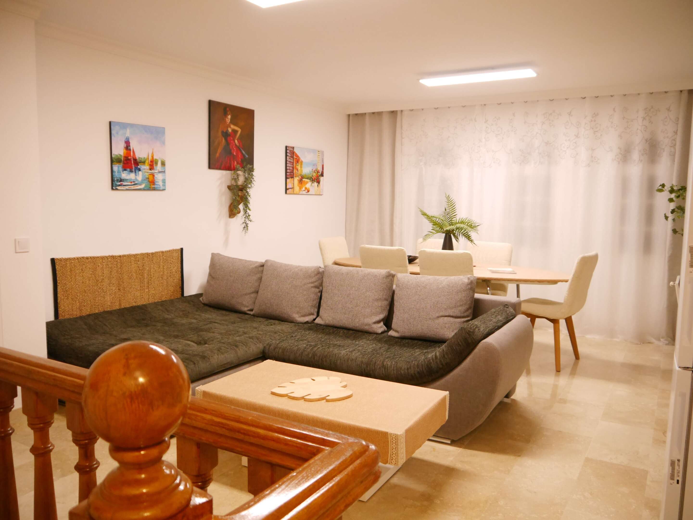 A cozy living room with a sofa, a wooden coffee table, and a dining area with a table and chairs. There are paintings on the wall and a plant on the dining table.
