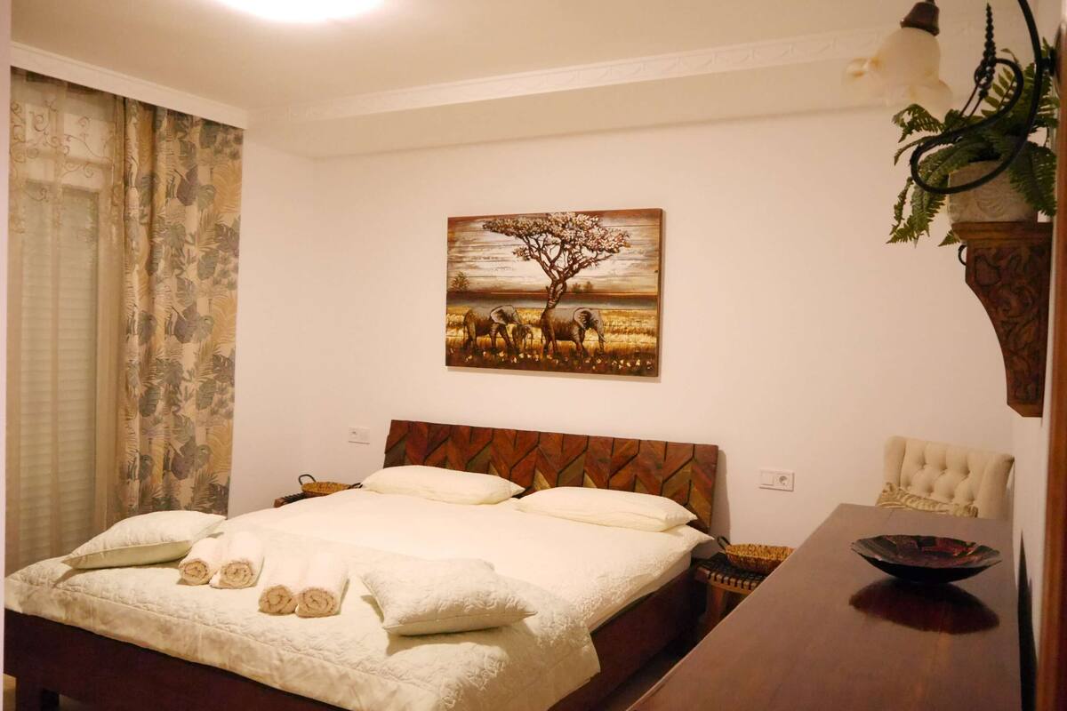 A bedroom with a bed, a painting of a tree and animals, a plant, and a table with a bowl.