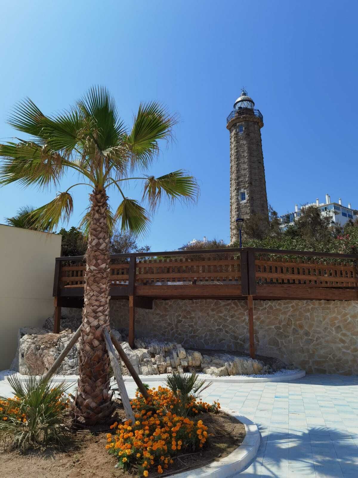 Una torre de piedra con techo metálico, palmeras y flores naranjas en un patio.