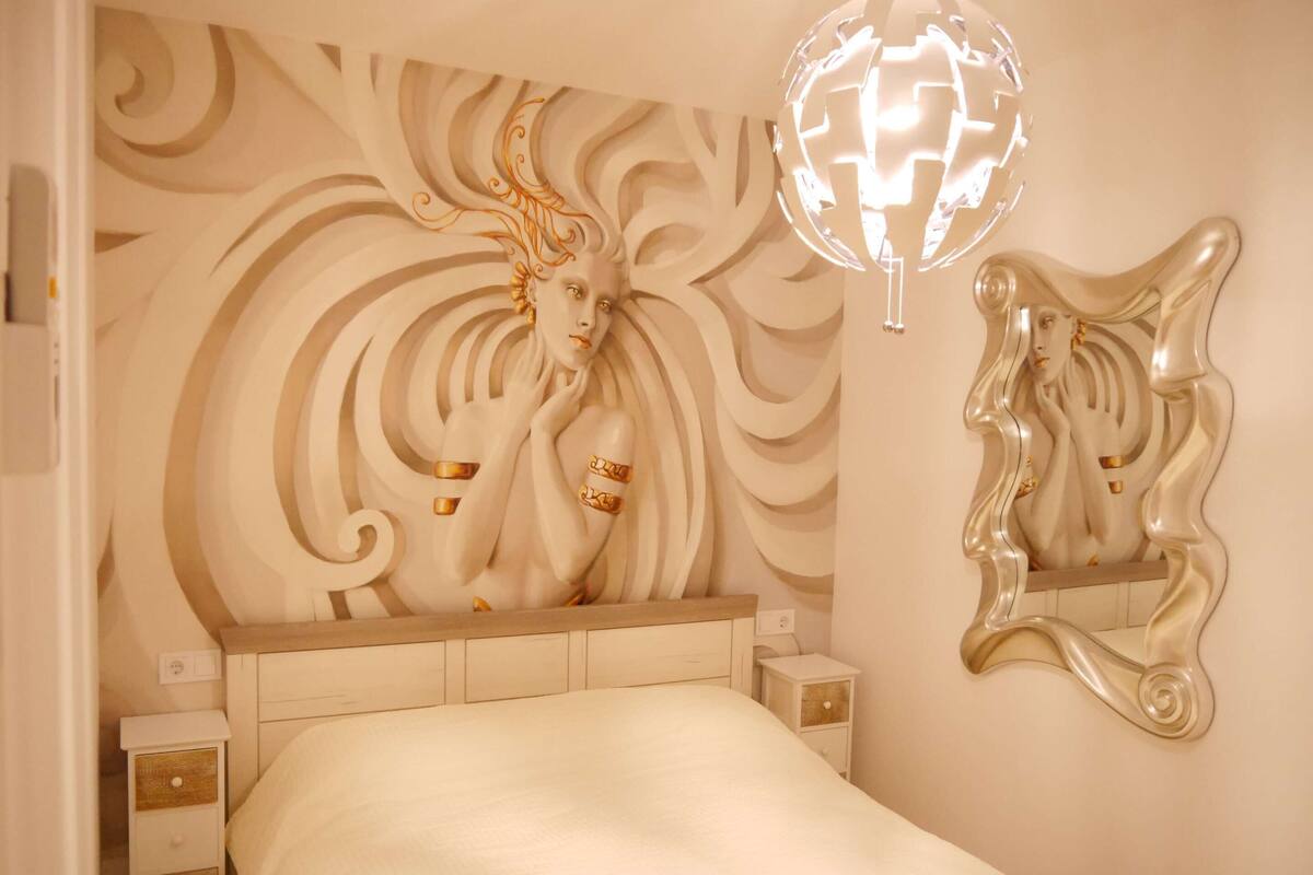 A bedroom with a large bed, a decorative headboard, a bedside table, and a wall-mounted mirror.