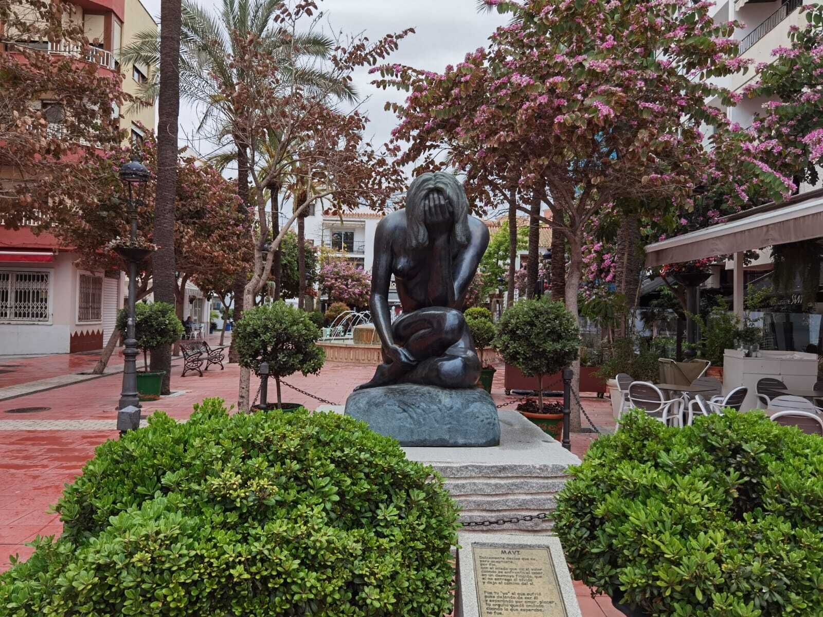 A statue of a seated figure in a public square with surrounding greenery and buildings.
