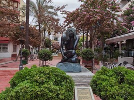 Estátua de uma figura sentada em praça pública, com áreas verdes e edifícios ao redor.