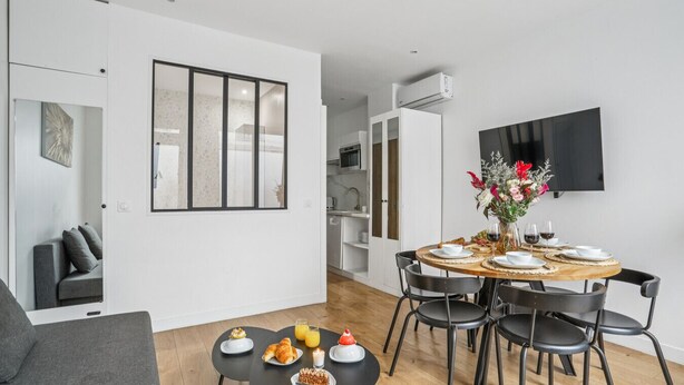 A modern living room with a dining area, a grey sofa, a flat-screen TV, and a dining table set with plates, glasses, and a vase of flowers.