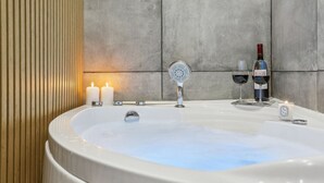 A modern bathroom with a freestanding bathtub, two lit candles, a bottle of wine, and a small candle on the edge of the tub.