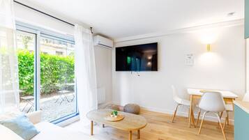 A modern living room with a large TV, a dining area, and a sliding glass door leading to a garden.