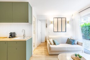 Una cocina moderna con un lavamanos, una cafetera y un mueble verde. Hay un sofá blanco con cojines y una pequeña mesa con plantas.