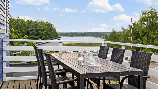 En træterrasse med et bord og stole, med udsigt over en sø omgivet af grønt.