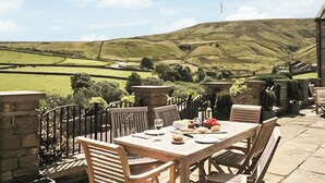 Un espace de restauration en plein air avec des murs en pierre, une table dressée pour deux personnes et une vue sur des collines verdoyantes en arrière-plan.