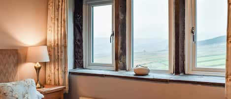 A bedroom with a bed, a nightstand, a lamp, a window with a view of the countryside, and a radiator.