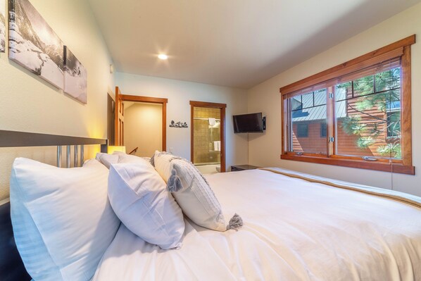 A bedroom with a large bed, a television mounted on the wall, and a window with a view of greenery outside.