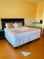 Una cama bien hecha con edredón blanco y manta beige, cabecera negra y una mesita de noche con hervidor y taza.
