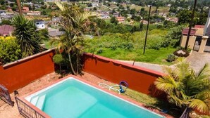 A rectangular swimming pool with a metal fence, surrounded by a red wall and greenery. - THE HAVEN GUEST HOUSE THEMBELIHLE  (Mbabane)