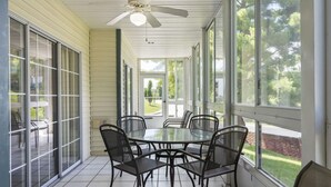 A covered patio with a glass table and chairs, large windows, and a ceiling fan.