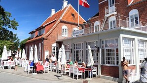 Un bâtiment traditionnel en briques, avec un toit recouvert de tuiles rouges, un drapeau et des places assises à l’extérieur, sous des parasols.