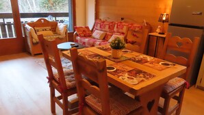 Un rincón comedor acogedor con una mesa de madera dispuesta para cuatro, rodeada de sillas a juego. Un sofá con cojines estampados se encuentra al fondo, y una mesita con una lámpara está a un lado.