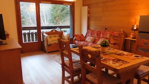 Una habitación acogedora con una mesa de madera, sillas y un sofá con cojines estampados. Una planta pequeña está sobre la mesa, y una lámpara sobre una mesita auxiliar.