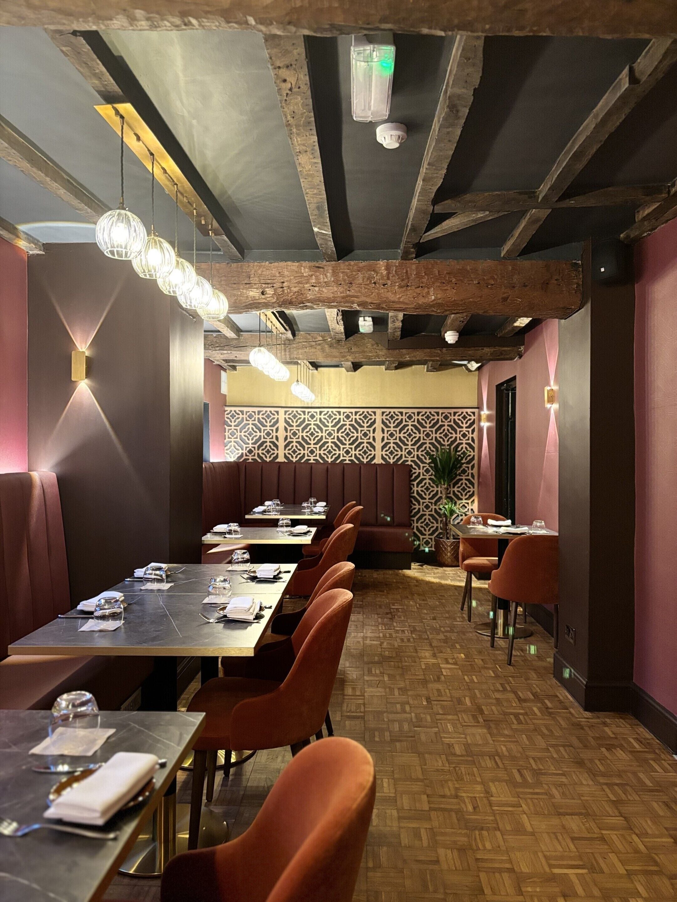Un restaurante con vigas de madera, papel tapiz estampado y sillas anaranjadas. Las mesas están preparadas con vasos y cubiertos.