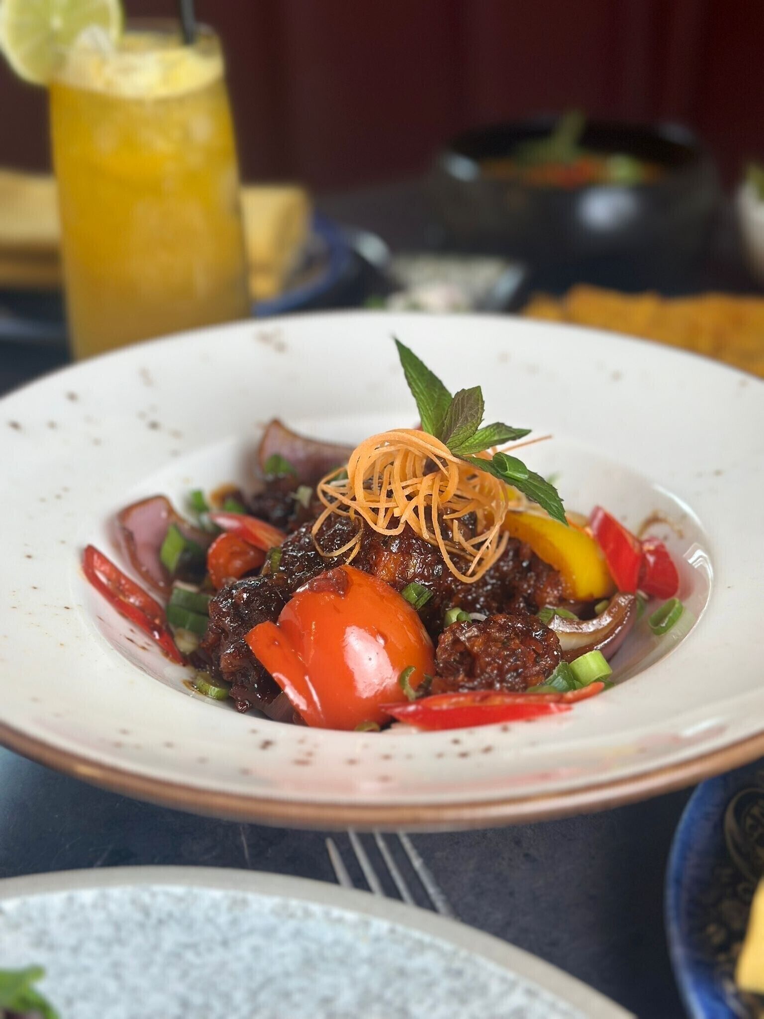 A plate of food with a garnish of mint leaves and noodles, accompanied by a drink in the background.