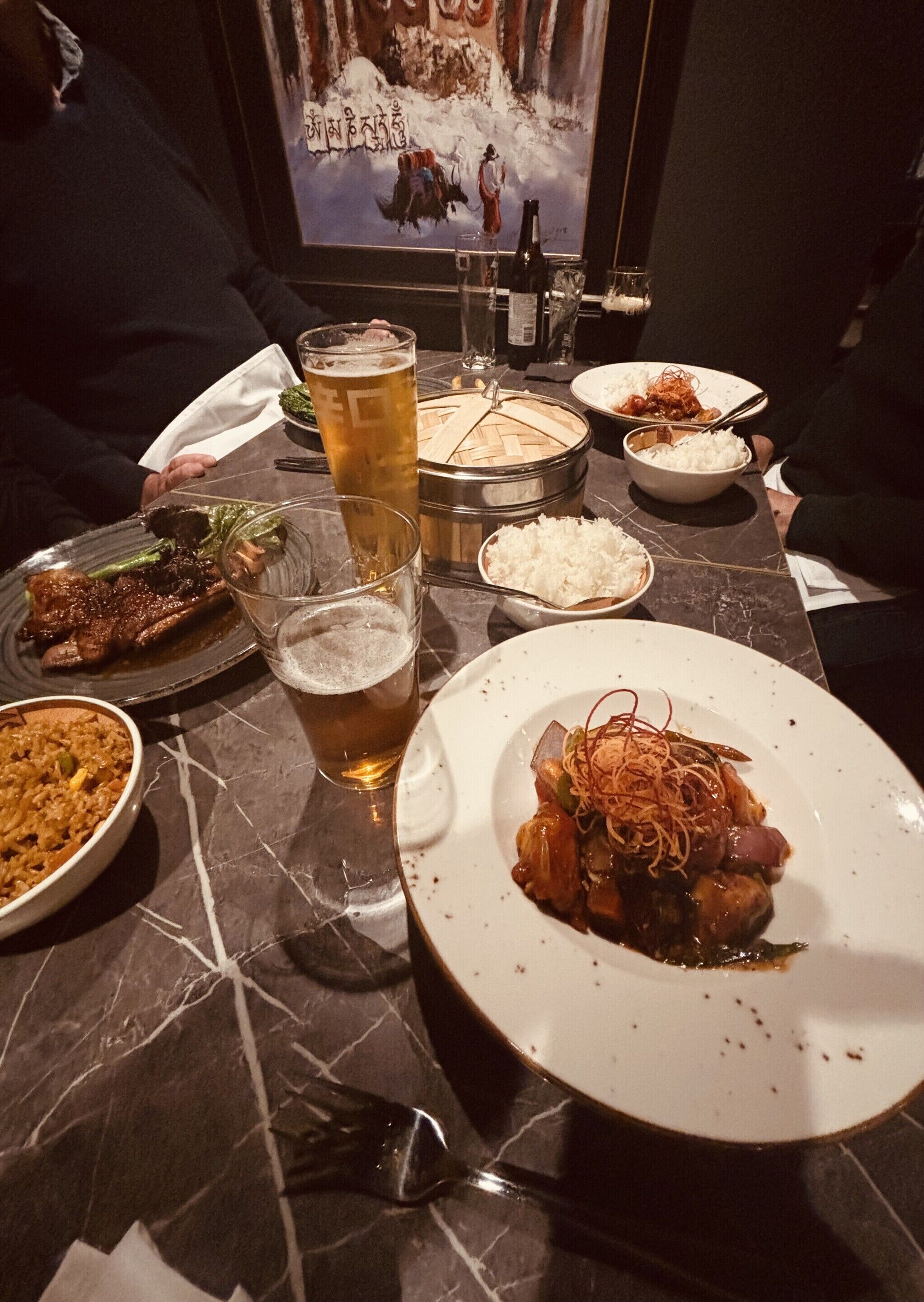 Una mesa de comedor con varios platos, incluyendo un tazón de arroz, un plato de vegetales salteados y un tazón de sopa. También hay varios vasos de cerveza y una botella de vino.