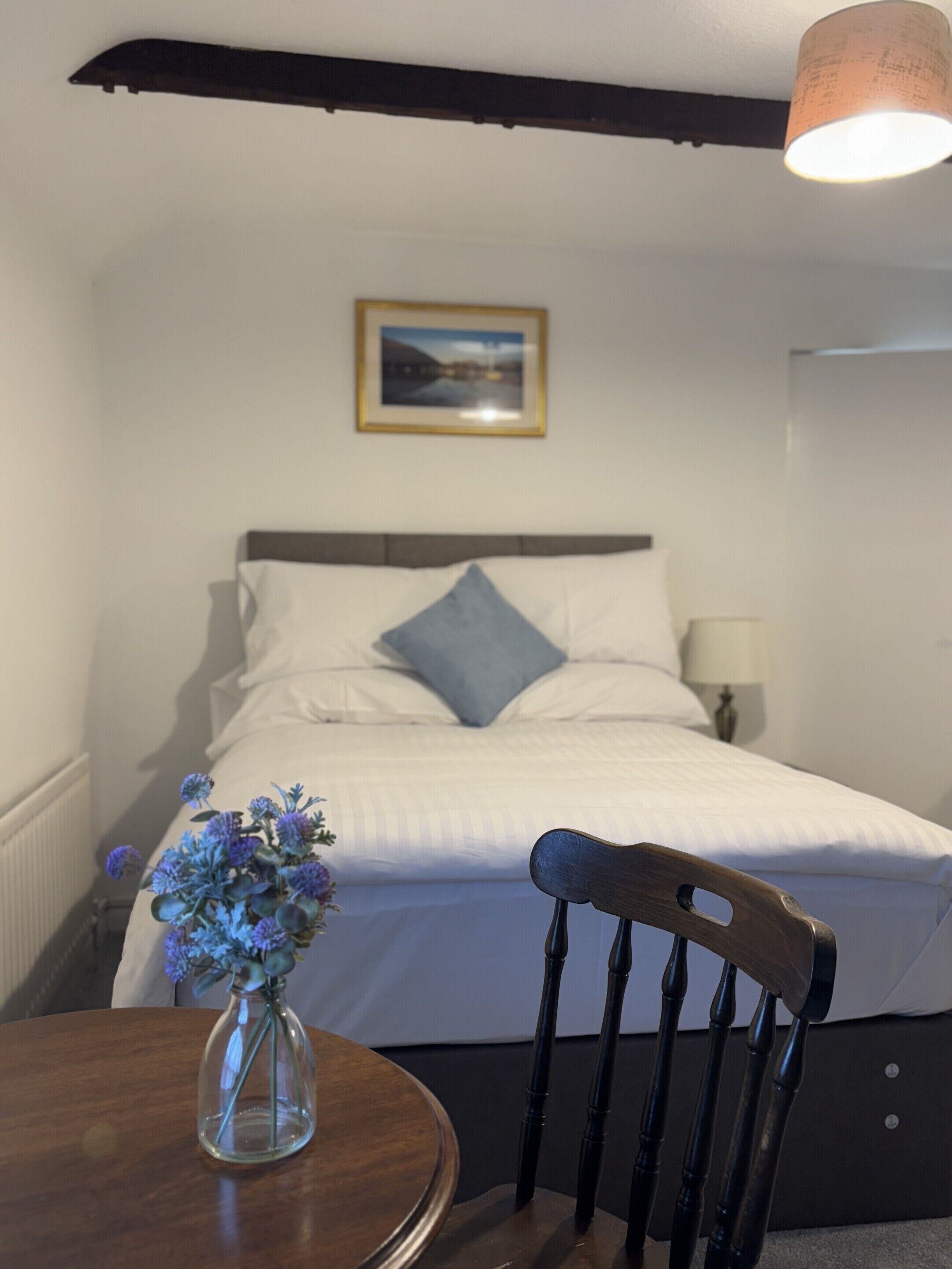 Um quarto com cama, mesa de cabeceira com um vaso de flores, cadeira, abajur e um quadro na parede.