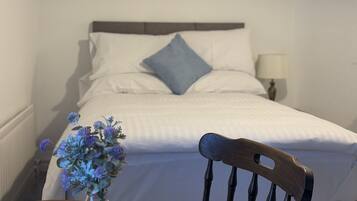 Um quarto com cama, mesa de cabeceira com um vaso de flores, cadeira, abajur e um quadro na parede.