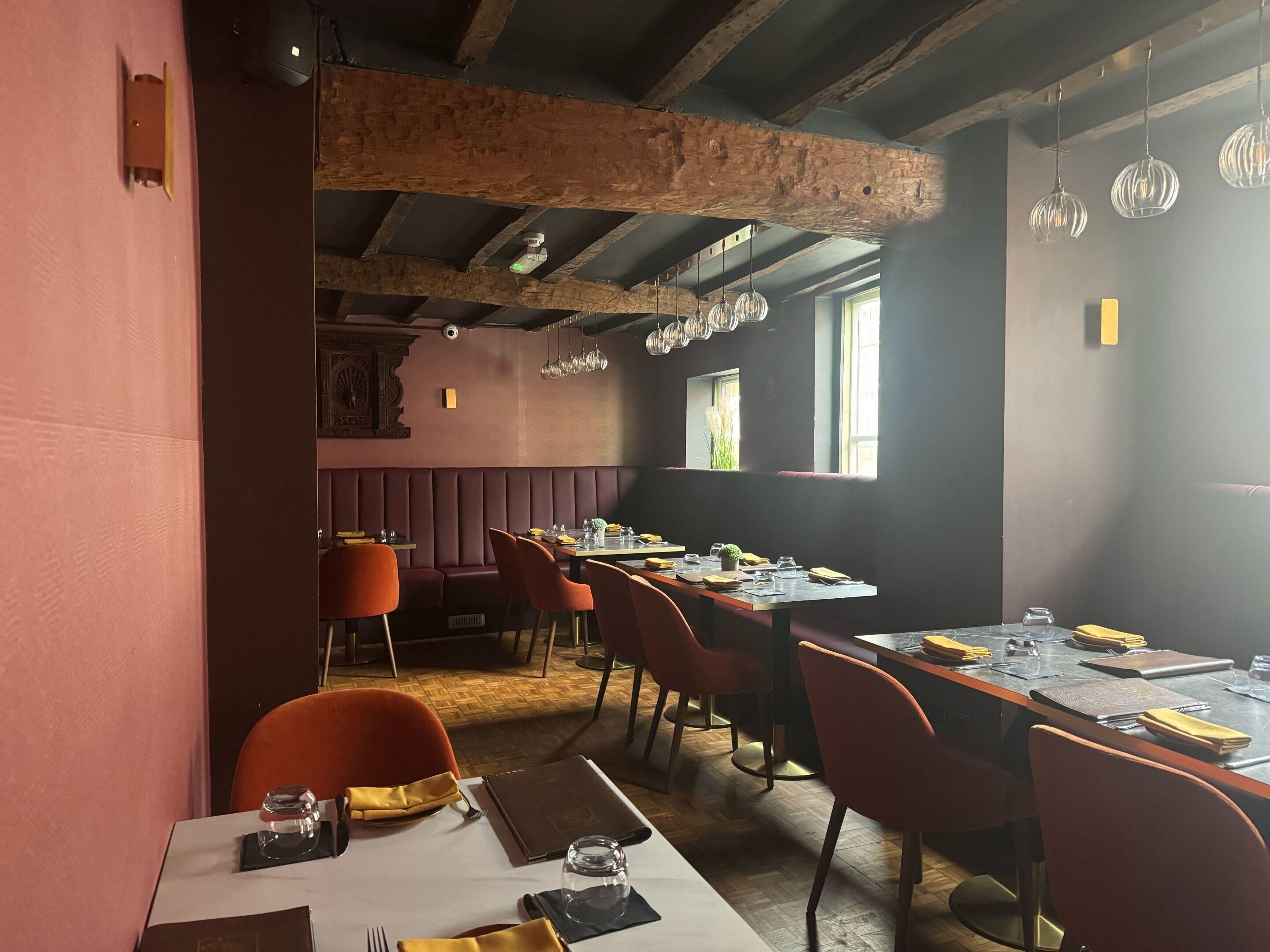 Ein Restaurant mit Holzbalken, roten Sitzbänken und orangefarbenen Stühlen. Die Tische sind mit Tellern, Gläsern und Servietten gedeckt.