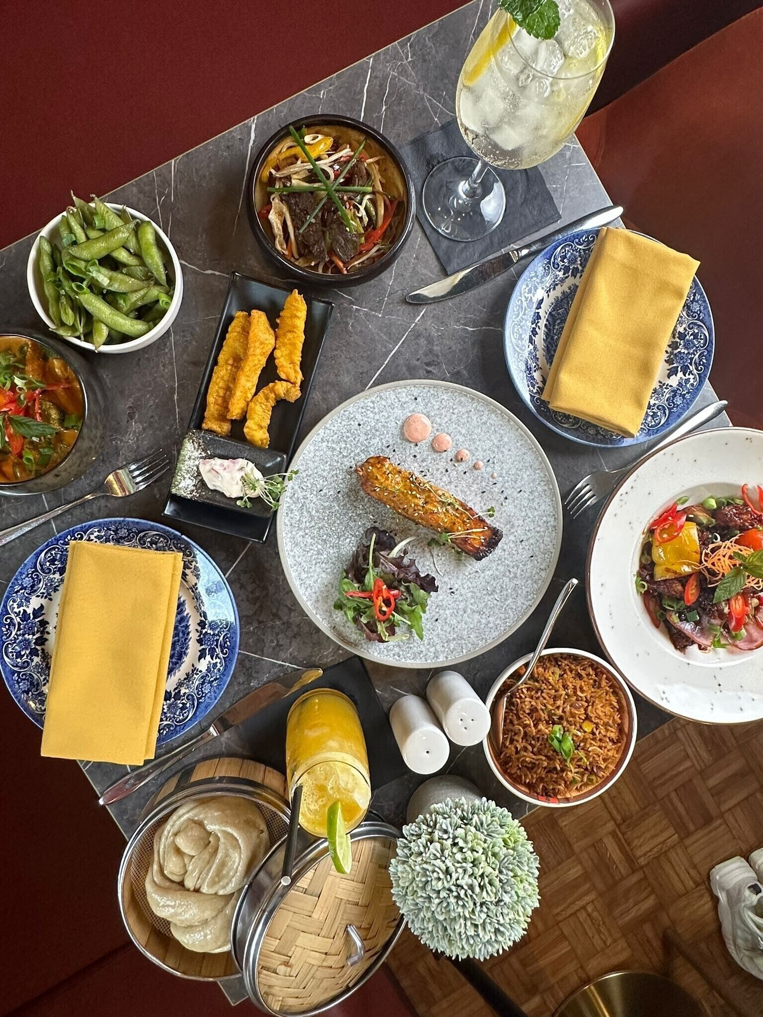 A table set with various dishes, including a bowl of green beans, a bowl of soup, fried items, a plate with a fish and salad, a plate with rice, and a glass of iced drink.