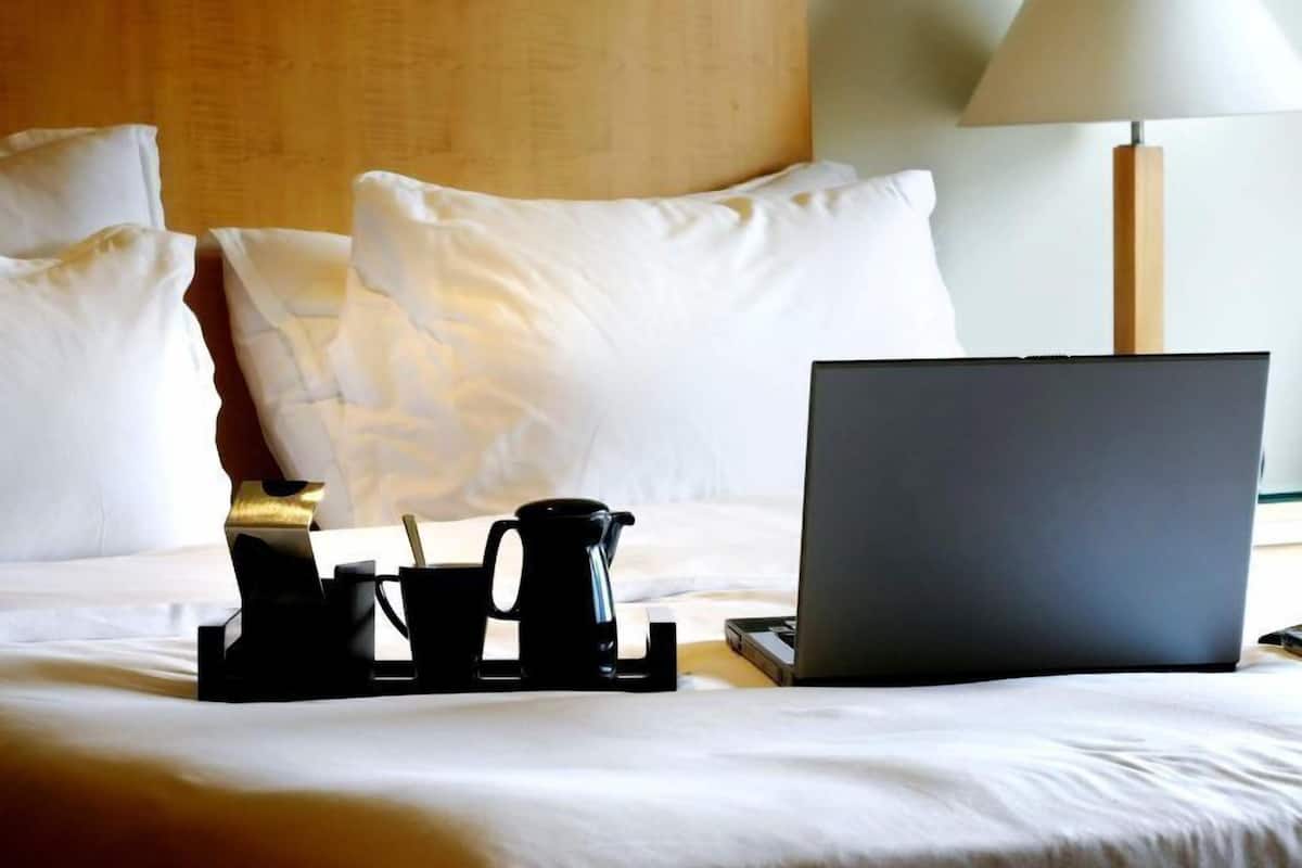 A neatly made bed with white linens, a laptop, a coffee maker, and a tray with cups and a spoon on a hotel room.