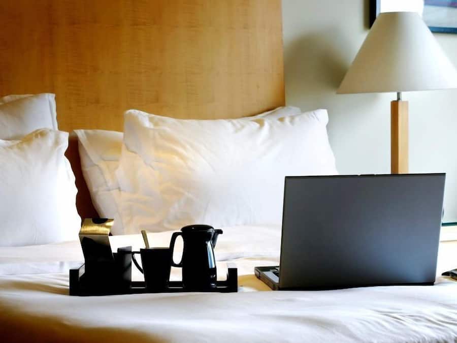 Una cama bien hecha con sábanas blancas, una computadora portátil, una cafetera y una bandeja con tazas y una cuchara en una habitación de hotel.