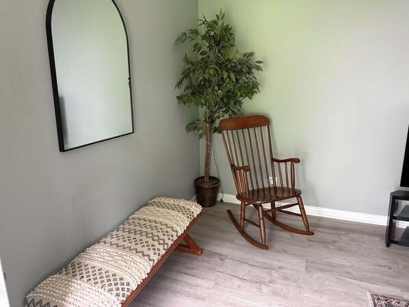 A room with a wooden rocking chair, a bench with a patterned cushion, a potted plant, and a wall-mounted mirror.