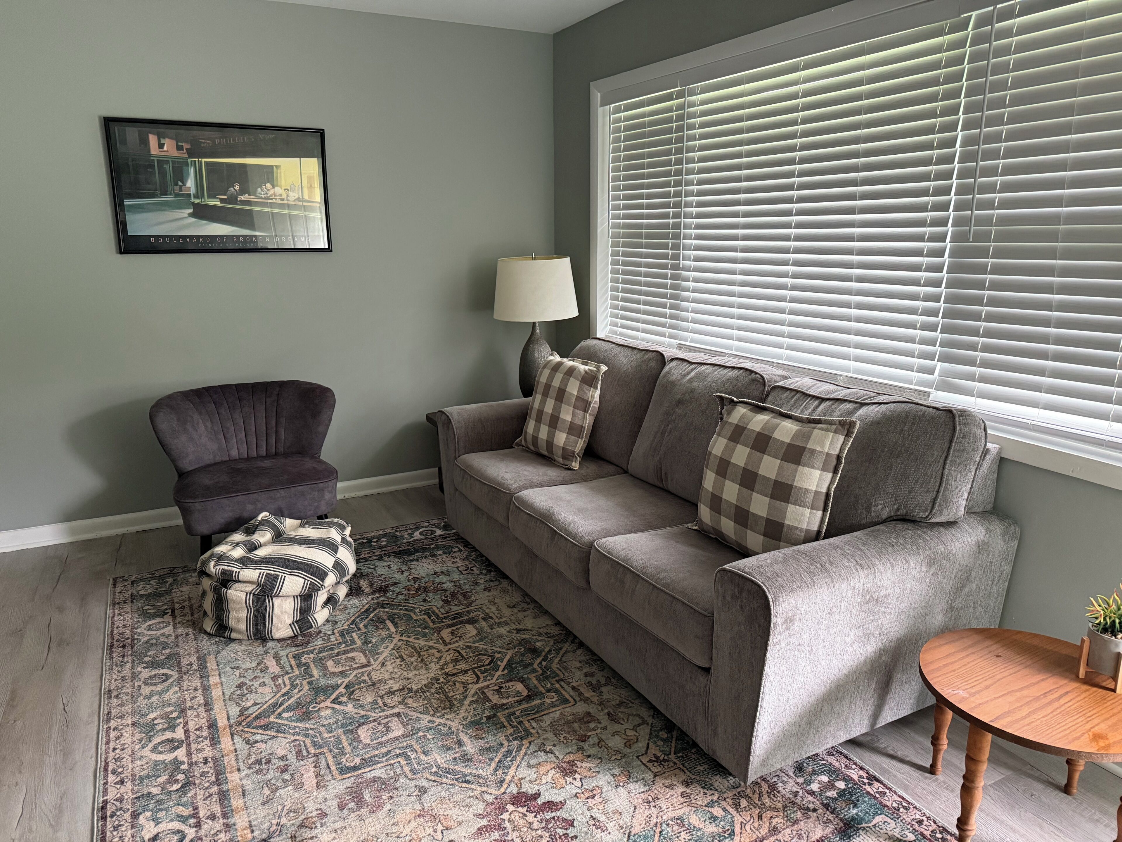 A living room with a grey sofa, a patterned rug, a small round wooden table, and a lamp.