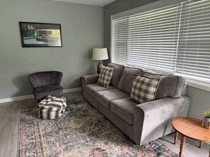 A living room with a grey sofa, a patterned rug, a small round wooden table, and a lamp.