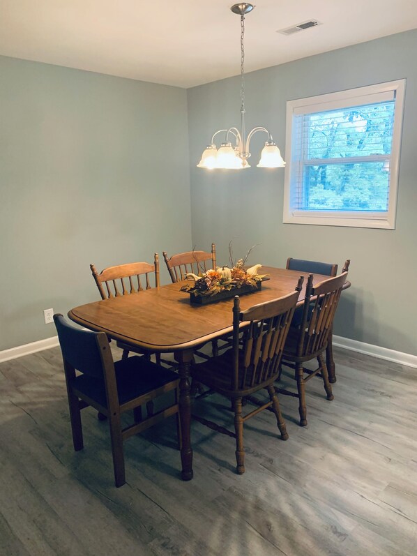 A dining area with a wooden table and chairs, a chandelier, and a window with blinds.