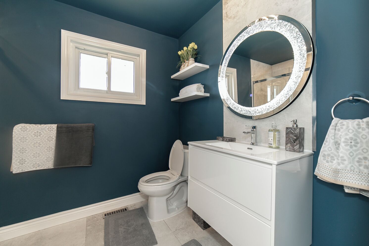A bathroom with a toilet, sink, and mirror. There is a window above the toilet and a towel rack on the wall.