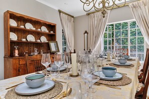 Ein Esszimmer mit einem langen Tisch, der für eine Mahlzeit gedeckt ist, einer Holzkolonne mit Geschirr und einem Fenster mit Vorhängen.