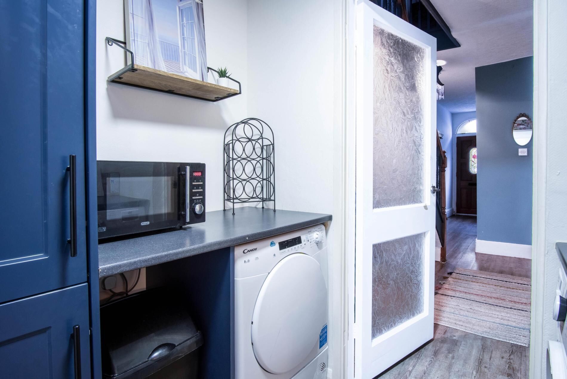 A compact kitchen with a microwave, a washing machine, and a small shelf with a plant.