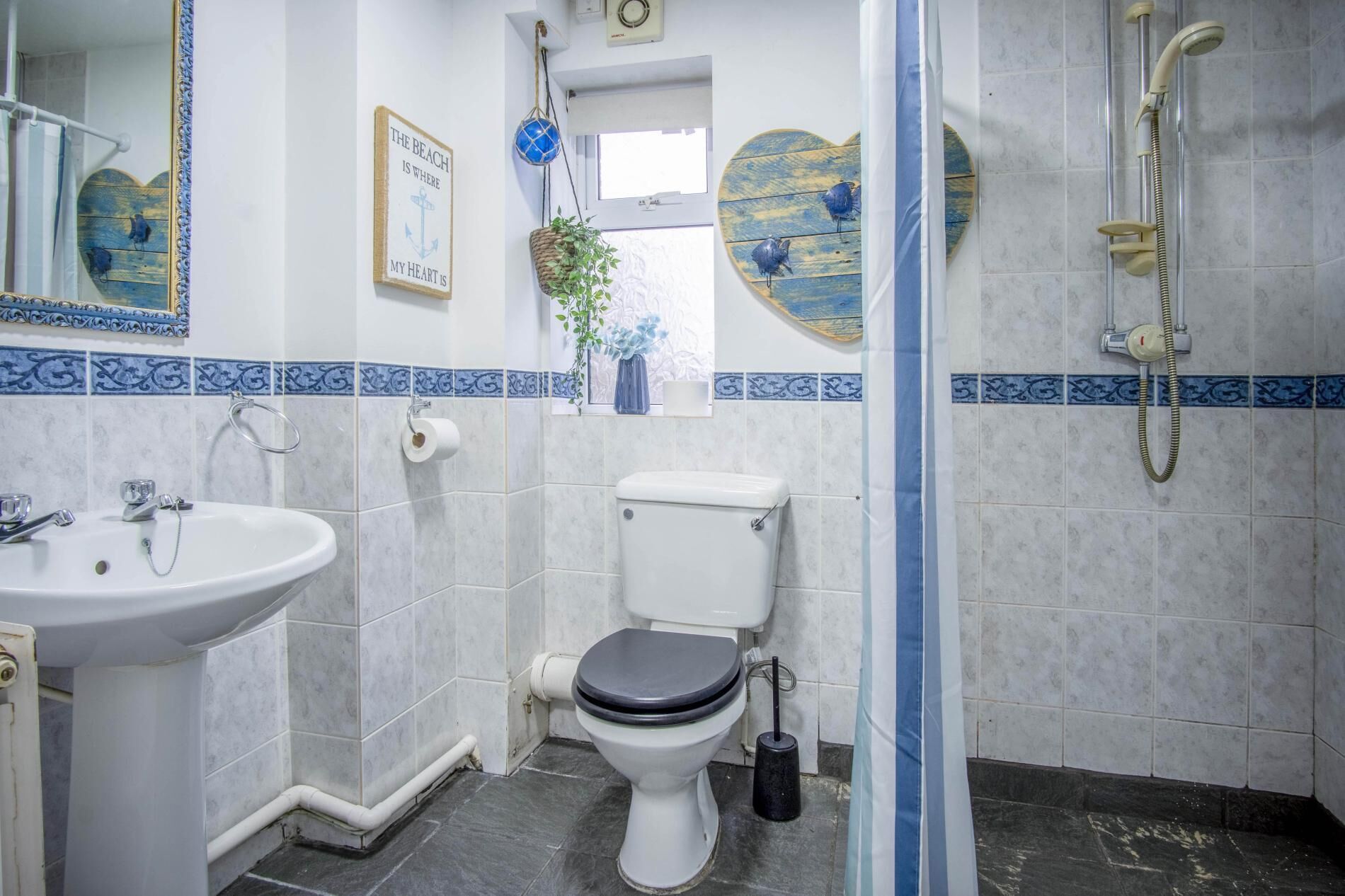 A bathroom with a white sink, toilet, and shower. There are decorative items like a heart-shaped mirror and a framed picture on the wall.
