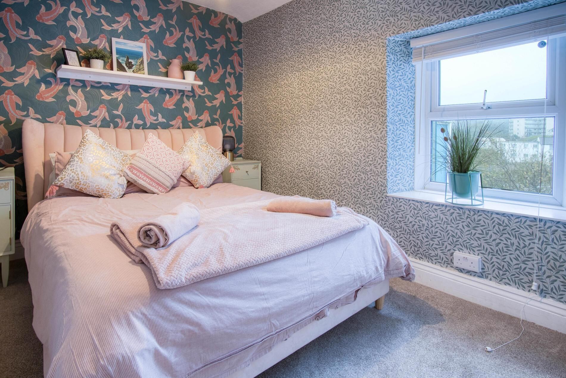 A bedroom with a bed, pillows, a nightstand, a window with a plant, and patterned wallpaper.